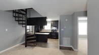 This two-story residence at The Fields offers a well-designed layout that balances functionality with contemporary style. The main level showcases an open-concept kitchen equipped with dark espresso cabinetry, black appliances including a range and refrigerator, and a kitchen island that provides additional workspace and casual dining potential. A window above the sink allows natural light to brighten the cooking area, while wood-look flooring runs throughout the kitchen, creating a warm, cohesive aesthetic.

The transition between the kitchen and adjacent living areas is seamless, with a combination of hard flooring and carpet defining distinct zones within the space. Neutral gray walls provide a versatile backdrop throughout, complementing the dark tones of the cabinetry and fixtures.

One of the most distinctive architectural features is the spiral staircase, finished in dark metal with wood-toned steps, which connects the main level to the upper floor. Its compact, elegant design makes an immediate visual statement while efficiently utilizing vertical space. The textured ceiling adds depth overhead, and thoughtful details such as a wall-mounted thermostat and floor-level vents reflect the home's practical, move-in-ready condition. Overall, the space presents a modern, coordinated interior suited for comfortable everyday living.