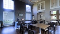 This single-floor space offers an impressive communal area designed for comfort and functionality. The room immediately draws attention with its soaring double-height ceilings, which create an open, airy atmosphere enhanced by large arched windows that flood the interior with natural light. A striking textured accent wall in a dark mosaic pattern adds a bold visual element, contrasting beautifully with the soft blue-gray walls throughout.

The space is furnished with multiple high-top and standard-height tables paired with dark leather-upholstered chairs, providing flexible seating arrangements suited for both individual use and small group gatherings. A built-in counter along one wall serves as a coffee and refreshment station, equipped with a coffee maker and beverage supplies, making it a convenient hub for daily use.

An elegant chandelier anchors the central area, adding a refined touch to the overall aesthetic. Additional cabinetry with glass-front doors is visible on the upper wall, providing both storage and decorative interest. Dark hardwood flooring runs throughout, grounding the space with warmth and continuity. Overall, the design balances sophistication with practicality.
