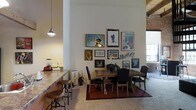 Located at 300 Blackwell St in the heart of Durham, NC, this two-story space blends industrial character with modern comfort. The interior showcases hallmark loft-style architecture, including exposed brick walls, soaring ceilings with visible structural elements, and generous windows that invite abundant natural light throughout.

The open-concept main level seamlessly integrates living and dining areas, creating a fluid and social atmosphere. The dining area serves as a distinct focal point, anchored by a solid wood table and complemented by a curated gallery wall that adds artistic personality to the space. Industrial-style pendant lighting casts a warm glow, enhancing the inviting ambiance.

Adjacent to the dining area, a kitchen counter with bar-height seating and round wooden stools provides a casual space for quick meals or conversation. The stone countertop and practical layout make the kitchen both functional and visually appealing.

Throughout the space, the interplay of raw materials — exposed brick, warm wood tones, and neutral carpeting — creates a layered, textured environment that feels both urban and welcoming. The two-floor configuration offers versatile use of space, with each level contributing to the overall sense of scale and openness. This property captures the spirit of Durham's vibrant, creative urban character.