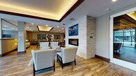 Located at 1709 Cresthill Ave in Royal Oak, Michigan, this single-floor communal lobby space is thoughtfully designed to balance comfort and functionality. The open-plan layout flows seamlessly across wood-look luxury vinyl flooring, connecting several distinct activity zones within one cohesive environment.

The lounge area serves as the centerpiece, anchored by a floor-to-ceiling stacked stone accent wall housing a linear gas fireplace beneath a wall-mounted flat-screen television. Cream-upholstered armchairs and a sofa are arranged around a low coffee table atop a plush area rug, creating an inviting gathering space. A tray ceiling with warm indirect cove lighting and dark wood trim frames this area elegantly.

Adjacent to the lounge, a casual dining zone offers multiple round tables paired with traditional wooden chairs, suitable for everyday socializing. High-top bistro tables along the wall provide additional seating beneath a curated gallery of circular and abstract artwork.

A practical service area includes package lockers, a multifunction printer, a small refrigerator, and counter space for resident use. Traditional mailboxes are also accessible within the space. Large glazed entry doors and expansive windows allow natural light to fill the interior while providing clear views to the surrounding streetscape, enhancing the open, welcoming ambiance throughout.