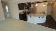This single-floor space showcases a well-designed open-concept layout where the kitchen flows naturally into the adjacent living area. The kitchen is outfitted with rich, dark espresso cabinetry that provides a striking contrast against the light granite countertops. Stainless steel appliances, including a range with microwave above and a full-size refrigerator, lend a modern, polished aesthetic to the cooking space. A curved breakfast bar with decorative corbel brackets extends from the kitchen, offering casual seating and serving as a natural dividing element between the kitchen and living spaces. Pendant lighting hangs above the bar, adding warm, focused illumination to the area, while recessed lighting throughout the kitchen ensures the workspace remains well-lit. The transition from the dark hardwood-style kitchen flooring to the plush, neutral-toned carpet of the living area creates a clear yet seamless distinction between the two functional zones. Crown molding along the upper walls adds a refined architectural detail, contributing to the overall clean and contemporary character of the space.