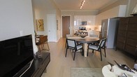 This single-floor residence in the United States showcases a thoughtfully designed open-concept layout that connects the living, dining, and kitchen areas into one cohesive and functional space. Wide-plank light hardwood flooring runs throughout, lending warmth and continuity to the interior. The kitchen is outfitted with sleek white cabinetry, stainless steel appliances including a range, microwave, and refrigerator, and is accented by track lighting that highlights the clean, modern aesthetic. Crown molding adds a refined architectural detail along the ceiling line, elevating the overall design.

The dining area features a round wood-topped table with stylish dark-framed chairs, comfortably accommodating multiple guests and serving as a natural centerpiece between the kitchen and living spaces. Adjacent to the dining area, the living space includes a media console and eclectic décor, including an eye-catching piece of wall art, contributing to the home's contemporary character. A corridor leads to additional rooms beyond, hinting at further private living spaces. The neutral color palette throughout creates a calm and inviting ambiance, while the open floor plan ensures excellent flow and a sense of spaciousness across the entire residence.