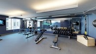 This single-floor fitness center is designed to support a comprehensive workout experience, offering a variety of exercise zones within a cohesive, modern interior. The space is defined by its bold navy blue walls, neutral gray carpet and rubber flooring, and bright recessed and linear lighting that creates a clean, energizing atmosphere throughout.

The cardio zone features a row of treadmills, elliptical machines, and stationary bikes positioned along floor-to-ceiling windows that overlook an outdoor courtyard, providing natural light and an open sense of space. Adjacent to this area, a dedicated strength training section houses a full rack of dumbbells, adjustable benches, and cable machines, complemented by a large wall-mounted mirror that enhances depth and visibility during workouts.

A designated training zone is equipped with a functional fitness wall system, including TRX straps, medicine balls, and resistance equipment, supporting dynamic and bodyweight training routines. A separate, open-plan room with carpeted flooring and a wall-mounted television serves as a flexible space suitable for stretching, yoga, or group fitness activities.

Throughout the fitness center, wall-mounted televisions provide entertainment, while the well-organized layout ensures ample room to move freely between equipment zones.