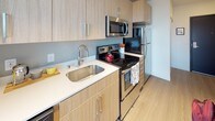This contemporary single-floor residence offers a thoughtfully designed open layout that maximizes both functionality and comfort. The kitchen is outfitted with light wood cabinetry, white countertops, a deep stainless steel sink, and a full suite of stainless steel appliances including an electric range, microwave, and refrigerator — providing an efficient and stylish cooking space. Light wood-style flooring flows consistently throughout the unit, lending warmth and visual continuity across all areas. The living area connects seamlessly to the kitchen, creating an inviting open-concept space. The primary bedroom is a standout feature, showcasing floor-to-ceiling sliding glass doors that open to a private balcony with expansive urban views of the surrounding cityscape. Exposed concrete ceiling details add an industrial character to the room. A second bedroom offers a cozy, well-organized layout with a built-in closet and dedicated desk area, ideal for work or study. The bathroom is clean and modern, featuring light wood-tone cabinetry, a toilet, and a shower-tub combo with a striped curtain, all set on wood-style flooring. The unit also includes an electrical panel, reflecting practical, move-in-ready infrastructure throughout.