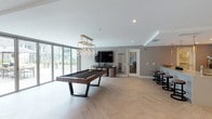 Located at 3425 W Olive Ave in Burbank, California, this contemporary community amenity space is designed to offer residents a stylish and versatile gathering environment. The single-floor layout is unified by elegant herringbone-patterned light wood flooring that flows throughout, lending the space a cohesive, upscale aesthetic.

The game room area serves as a dynamic focal point, anchored by a sleek billiards table set beneath a gold-toned globe pendant chandelier. A wall-mounted flat-screen TV and cue rack complete the entertainment setup, while large floor-to-ceiling sliding glass doors open directly to an outdoor terrace, flooding the space with natural light and providing seamless indoor-outdoor access.

Adjacent to the game area, the lounge zone offers a warm, inviting retreat featuring a tufted sofa, rounded armchairs, and gold accent tables arranged atop a patterned area rug. A linear gas fireplace set against a stacked-stone accent wall adds a sophisticated ambiance, while a built-in media console and sheer floor-to-ceiling window treatments enhance the refined atmosphere. A private booth nook with padded banquette seating offers an intimate setting for smaller gatherings.

The catering kitchen is well-appointed with light wood cabinetry, quartz countertops, a stainless steel refrigerator, and a striking stacked-stone accent wall, making it ideal for hosting events.