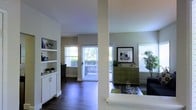 This single-floor residence offers an open and airy layout designed for comfortable everyday living. The main living area is anchored by rich, dark hardwood flooring that extends throughout, creating a warm and cohesive aesthetic. A structural column subtly defines the transition between the living and dining zones while preserving the open-plan feel.

Natural light is abundant, streaming in through multiple large windows fitted with horizontal blinds, as well as a sliding glass door that opens directly onto a private balcony. This indoor-outdoor connection enhances the sense of space and brings in refreshing views of surrounding greenery.

Built-in white shelving units provide ample display and storage space, complemented by white cabinetry below — a practical yet clean design detail. The living area is well-proportioned, accommodating seating and accent furniture with ease. A decorative green dresser and framed wall art add personality and a pop of color against the neutral, softly toned walls.

The overall ambiance is light, modern, and welcoming, with a neutral color palette of whites, greens, and warm wood tones that creates a calm and livable environment throughout the space.
