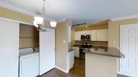 This single-floor space offers a well-organized, open layout that seamlessly connects the kitchen and adjacent living areas. The kitchen is a standout feature, equipped with stainless steel appliances including an electric range, built-in microwave, and a double-basin sink. Dark granite countertops provide ample workspace and extend to a peninsula that serves as a natural divider between the kitchen and the dining or living area. White cabinetry lines the upper and lower walls, offering generous storage throughout. Track lighting above the kitchen ensures focused, functional illumination, while a stylish chandelier overhead adds a decorative touch to the adjacent space.

The warm, golden-toned walls complement the rich, dark hardwood flooring that runs throughout, creating a cohesive and inviting atmosphere. Crown molding along the ceiling adds a refined architectural detail. A textured ceiling is consistent across the space. A particularly practical feature is the in-unit laundry area, discreetly concealed behind large sliding doors, housing a full-sized washer and dryer alongside additional shelving for storage. The overall layout is efficient, maximizing functionality within a compact footprint.