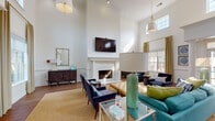 This single-story leasing center in Collierville, Tennessee is designed to make a strong first impression through a combination of sophisticated architectural details and thoughtfully curated interiors. The space is defined by dramatically tall ceilings that lend an open, airy quality throughout, complemented by abundant natural light streaming in through large windows dressed with gold drapes and layered roller shades.

The main lounge area serves as the centerpiece of the space, anchored by a stately white-painted fireplace with a wall-mounted flat-screen television above it. Seating arrangements feature a vibrant teal sofa, navy blue accent chairs, and a tufted ottoman, all positioned atop a woven area rug over warm wood-look flooring. Pendant lighting with brass accents and oversized table lamps add warmth and dimension.

A dedicated office area provides a more functional setting, with a clean white desk, task seating, and views of the outdoors through generous windows. Private offices accessible via French doors offer additional workspace with a polished, professional feel.

A separate seating nook features a round marble-top table with wishbone chairs, ideal for one-on-one consultations. Decorative columns and wainscoting throughout reinforce the refined, classic aesthetic of this well-appointed leasing center.