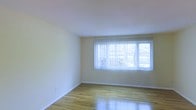 This single-floor space presents a straightforward and functional layout with a clean, neutral aesthetic throughout. The main living area is finished with light-toned hardwood flooring that reflects natural light, lending the room a bright and open feel. The walls are painted in a soft, off-white hue, creating a versatile backdrop that complements a variety of interior styles.

A large window fitted with vertical blinds serves as a focal point in the living area, allowing generous amounts of natural light to flow into the space while offering views of the surrounding outdoor environment. The window's size and placement contribute to an airy atmosphere within the room.

White baseboards run along the perimeter of the room, providing clean contrast against both the flooring and walls. The ceiling is smooth and bright white, further enhancing the sense of openness. Overall, the space maintains a minimalist character, keeping the focus on its functional features and the natural light that defines its ambiance. The single-floor layout offers ease of accessibility and a straightforward flow throughout.