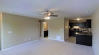 This single-floor residence offers a practical and comfortable layout designed for everyday living. The main living area is generously sized, finished with soft wall-to-wall carpeting in a neutral tone that creates a warm and inviting atmosphere. A ceiling fan with an integrated light fixture is centrally mounted, providing both air circulation and ambient lighting to the space. The walls are painted in a muted beige, lending the interior a clean and understated aesthetic.

Adjacent to the living area, the kitchen is clearly defined by a transition to tile flooring. It features dark, espresso-toned cabinetry that contrasts with the lighter surroundings, paired with black appliances including a range and refrigerator. The bold cabinetry gives the kitchen a sharp, classic character.

A doorway visible from the main living area leads to a bathroom, which offers dark vanity cabinetry consistent with the kitchen's design language, maintaining a cohesive look throughout the unit. The layout connects the rooms efficiently, making the overall flow of the space feel intuitive and functional. With its neutral finishes and thoughtful design, this single-level residence offers a solid foundation for comfortable everyday living.