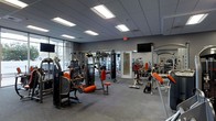 This fitness center offers a comprehensive workout environment designed to accommodate a wide range of training needs. The space is outfitted with a full array of professional-grade strength training machines finished in a striking black-and-orange color scheme, providing dedicated stations for targeting major muscle groups. A free weight section lines one wall, complete with a tiered dumbbell rack and adjustable benches, catering to both beginners and experienced lifters. Large floor-to-ceiling windows along one side of the facility flood the space with abundant natural light, creating an energizing atmosphere. The interior is finished with a drop-tile ceiling fitted with recessed panel lighting, ensuring consistent illumination throughout the workout floor. Durable, textured carpet flooring in a neutral gray tone provides both comfort and traction underfoot. Wall-mounted flat-screen televisions are positioned at multiple points around the room, offering entertainment during workouts. Full-length mirrors along one wall enhance the sense of openness while allowing members to monitor their form. The layout is thoughtfully organized to allow ample space between equipment, ensuring a safe and comfortable training experience. The overall design is clean, modern, and functional, making it a well-rounded facility for fitness enthusiasts.