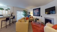 This thoughtfully designed single-floor residence offers a comfortable and functional layout across several well-defined living spaces. The living room serves as the heart of the home, featuring a classic fireplace flanked by built-in speakers, a wall-mounted television, and sliding glass doors that invite natural light and provide outdoor access. Warm tones, bold artwork, and lush indoor plants contribute to an inviting ambiance throughout.

The kitchen is efficiently arranged with ample white cabinetry accented by wood trim, generous counter space, and full appliance suite including a dishwasher, range, and microwave. A versatile den or home office adjacent to the main living area offers a bright, functional workspace complete with a glass-top desk, storage shelving, and a vibrant red sofa, easily adaptable to various needs.

The primary bedroom accommodates a dramatic four-poster bed and benefits from multiple windows with outdoor views. A second bedroom mirrors this layout. Both bathrooms are finished with light tile flooring; the primary bath features a wide vanity with dark countertops and a large mirror, while the secondary bath includes a shower-tub combination with decorative curtains. Throughout, neutral walls and carpeted bedrooms provide a calm, cohesive backdrop.