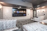Located at 1669 Scott Blvd in Decatur, Georgia, this single-level private screening and lounge space is thoughtfully designed to deliver an upscale, cinema-inspired experience. The room is anchored by a large wall-mounted display screen and outfitted with multiple tiers of plush, oversized sectional seating upholstered in a sophisticated silver-toned jacquard fabric, accented with colorful round throw pillows and paired with sleek mirrored and gold side tables. The walls are clad in a distinctive geometric-patterned wallcovering in soft champagne and taupe tones, which lends a modern yet refined texture throughout the space. A series of elegant black iron wall sconces lines the walls, casting warm, intimate ambient light that complements the dark, dramatic ceiling treatment. Patterned area carpeting in deep charcoal and grey tones grounds the interior and reinforces the lounge atmosphere. Recessed lighting provides additional illumination, while smart control panels are conveniently integrated into the walls. A private entry door with a dark finish ensures a discreet, contained environment. The overall ambiance is intimate and luxurious, making this space well-suited for private screenings, group entertainment, or exclusive social gatherings.