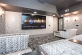 Located at 1669 Scott Blvd in Decatur, Georgia, this single-level private screening and lounge space is thoughtfully designed to deliver an upscale, cinema-inspired experience. The room is anchored by a large wall-mounted display screen and outfitted with multiple tiers of plush, oversized sectional seating upholstered in a sophisticated silver-toned jacquard fabric, accented with colorful round throw pillows and paired with sleek mirrored and gold side tables. The walls are clad in a distinctive geometric-patterned wallcovering in soft champagne and taupe tones, which lends a modern yet refined texture throughout the space. A series of elegant black iron wall sconces lines the walls, casting warm, intimate ambient light that complements the dark, dramatic ceiling treatment. Patterned area carpeting in deep charcoal and grey tones grounds the interior and reinforces the lounge atmosphere. Recessed lighting provides additional illumination, while smart control panels are conveniently integrated into the walls. A private entry door with a dark finish ensures a discreet, contained environment. The overall ambiance is intimate and luxurious, making this space well-suited for private screenings, group entertainment, or exclusive social gatherings.