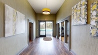 This single-story amenity building offers a compact yet functional layout comprising a welcoming hallway and a dedicated exercise room. The hallway sets an inviting tone with warm hardwood-style flooring, textured olive-green walls, and a stylish drum pendant light. Glass-paneled doors line both sides, providing direct sightlines into the fitness area and creating an open, connected feel. A glass exit door at the end of the corridor frames a view of the outdoor pool beyond, flooding the space with natural light.

The exercise room is well-equipped with multiple treadmills, an elliptical, a stair climber, a full dumbbell rack with bench, resistance bands, and a TRX suspension system. Light hardwood flooring, full-length mirrors, recessed lighting, and clerestory windows contribute to an airy, energizing atmosphere. A direct door provides convenient pool-side access.

Outside, the expansive pool deck is lined with rows of lounge chairs and side tables, ideal for relaxing in the sun. Manicured hedges and mature trees border the area, offering privacy and a pleasant natural backdrop. The surrounding residential buildings are visible in the distance, situating this facility as a central community amenity.