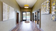 This single-story amenity building offers a compact yet functional layout comprising a welcoming hallway and a dedicated exercise room. The hallway sets an inviting tone with warm hardwood-style flooring, textured olive-green walls, and a stylish drum pendant light. Glass-paneled doors line both sides, providing direct sightlines into the fitness area and creating an open, connected feel. A glass exit door at the end of the corridor frames a view of the outdoor pool beyond, flooding the space with natural light.

The exercise room is well-equipped with multiple treadmills, an elliptical, a stair climber, a full dumbbell rack with bench, resistance bands, and a TRX suspension system. Light hardwood flooring, full-length mirrors, recessed lighting, and clerestory windows contribute to an airy, energizing atmosphere. A direct door provides convenient pool-side access.

Outside, the expansive pool deck is lined with rows of lounge chairs and side tables, ideal for relaxing in the sun. Manicured hedges and mature trees border the area, offering privacy and a pleasant natural backdrop. The surrounding residential buildings are visible in the distance, situating this facility as a central community amenity.