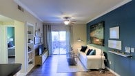 This single-floor space offers a thoughtfully designed living area that balances style and functionality. The living room serves as the centerpiece, featuring rich hardwood-style flooring, crown molding, and a ceiling fan that enhances air circulation and adds a classic architectural touch. A large sliding glass door floods the room with natural light and provides direct access to a private balcony, offering a pleasant outdoor retreat with views of surrounding greenery. The living room is anchored by a crisp white sofa accented with teal and navy throw pillows, complemented by a bold teal accent wall adorned with a striking piece of abstract artwork. Built-in open shelving on the opposite wall provides display and storage space alongside a media console with a wall-mounted television. Sheer chevron-patterned curtains frame the sliding door, softening the natural light while maintaining an airy feel. Coordinating floor lamps on either side of the seating area provide warm ambient lighting. The overall ambiance is modern yet welcoming, with a cohesive color palette of warm neutrals, deep teal, and natural wood tones that creates a visually harmonious and comfortable environment throughout the space.