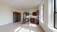 Located at 201 CTH-125 in Appleton, Wisconsin, this single-floor unit offers a thoughtfully designed layout with a blend of modern finishes and functional living spaces. The open-concept kitchen and living area serves as the heart of the unit, featuring rich dark wood cabinetry, stainless steel appliances including a refrigerator, microwave, and range, a subway tile backsplash, and light hardwood-style flooring that runs throughout. A rustic wood work table adds character to the kitchen space. Large, tall windows with clean white trim flood the area with abundant natural light and provide views of the surrounding Appleton streetscape. An arched doorway adds an architectural detail that connects the kitchen to the adjacent spaces. The open living area offers generous room to configure furnishings flexibly. The bedrooms are finished with soft gray carpeting and feature tall, narrow windows that continue the theme of natural light throughout the unit. The bathroom is cleanly appointed with tile flooring, a combination tub and shower with built-in shelving, a dark wood vanity with a light countertop, and recessed lighting. The unit also includes access to an exterior door, providing a private entry point. Recessed lighting and neutral wall tones throughout contribute to a clean, contemporary ambiance.