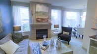 This thoughtfully designed single-floor residence offers a seamless blend of comfort and contemporary style. The living area serves as the heart of the home, anchored by a classic tiled fireplace with a clean-lined mantel that provides a natural focal point for the room. Framed by large windows on multiple walls, the space is bathed in abundant natural light that highlights the neutral, soft gray tones of the walls and the warm, textured flooring underfoot. The windows are dressed with flowing white curtains that enhance the airy, open ambiance while offering views of the lush greenery outside. The living area is elegantly appointed with a tufted sofa, a streamlined wingback accent chair, and a metallic coffee table adorned with curated decorative accessories. A large landscape painting above the fireplace adds a sophisticated artistic touch. The dining area is partially visible adjacent to the living space, suggesting an open and connected floor plan that encourages easy movement throughout the home. Overall, the space exudes a calm, polished atmosphere with a carefully curated mix of textures and finishes that make it both stylish and inviting.