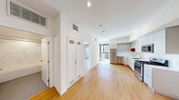 This contemporary single-floor unit showcases a clean, modern aesthetic throughout, characterized by crisp white walls, warm light hardwood flooring, and abundant natural light. The open-concept layout connects the living area seamlessly to the kitchen, which is well-appointed with sleek grey cabinetry extending to the ceiling, stainless steel appliances including a gas range, microwave, refrigerator, and dishwasher, as well as a functional sink with ample counter space. A distinctive built-in storage unit with louvered doors provides practical organization near the entryway. The bedroom is generously proportioned and features large floor-to-ceiling sliding glass doors that open directly onto a private balcony, offering open urban views of the surrounding cityscape. A second window in the bedroom further enhances the flow of natural light. The bathroom is elegantly finished with large-format marble-style wall tiles in the tub and shower surround, classic hexagonal floor tiling, a warm wood-toned floating vanity, and recessed lighting. Recessed ceiling lights throughout the unit complement the modern design, while centralized HVAC vents ensure year-round comfort. The balcony, accessible from the bedroom, provides an inviting outdoor retreat with views of neighboring architecture.