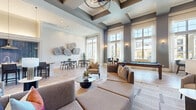 This elegantly designed single-floor community amenity space offers a generous and versatile layout ideal for socializing and recreation. The main lounge area commands immediate attention with its dramatic coffered ceilings accented in deep charcoal tones, complemented by a striking chandelier and recessed lighting that create a warm, inviting ambiance. Tall, floor-to-ceiling windows line the walls, flooding the interior with abundant natural light and offering views of the surrounding community.

The lounge is furnished with plush neutral sofas and accent chairs arranged around low coffee tables, creating intimate conversation areas. A full-size billiards table anchors the recreational zone, while multiple dining tables and chairs provide additional gathering space. Herringbone-patterned flooring in a light wood tone flows throughout, adding texture and visual warmth.

A sleek linear fireplace clad in large-format grey tile serves as a dramatic focal point in one area, framed by rich wood-and-glass double doors that elegantly partition sections of the space. The fully equipped kitchen features bold cobalt blue tile backsplash, navy cabinetry, granite countertops, stainless steel appliances, and a spacious island with bar seating. Tasteful wall art and sculptural elements complete the refined, contemporary aesthetic throughout.