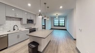 Located at 16 Carlson Ct in Charleston, SC, this single-floor residence presents a cohesive modern aesthetic throughout its open layout. The kitchen serves as a central highlight, outfitted with high-gloss gray cabinetry extending to the ceiling, quartz countertops, a white subway tile backsplash, stainless steel appliances including a French door refrigerator and dishwasher, and a central island with pendant lighting above. Light wood-look flooring runs throughout the main living areas, complementing the clean, neutral color palette.

The open-concept design connects the kitchen seamlessly to the living area, which is anchored by large, bold black-framed grid windows that invite generous natural light and offer elevated views of the surrounding Charleston cityscape and neighboring brick architecture.

The bedrooms continue the light flooring and neutral tones, each featuring large grid windows that reinforce the industrial-modern character of the space. The bathrooms are finished with dark large-format floor tiles contrasting with bright white walls and marble-style tile surround in the tub and shower. The primary bathroom features an extended vanity with dual sinks, ample drawer storage, and a wide mirror with overhead lighting. Gray panel doors throughout tie the interior together with a refined, consistent design language.