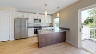 Located at 16 Farmstead Rd in Yarmouth, Maine, this single-floor residence offers a thoughtfully designed layout with modern finishes throughout. The kitchen serves as a central highlight, featuring crisp white cabinetry, granite countertops, a large dark-wood island with a built-in sink and pendant lighting above, and a full suite of stainless steel appliances including a refrigerator, range, and microwave. Light wood-style flooring flows through the main living areas, enhancing the bright, open feel of the space. A sliding glass door in the kitchen area opens to an outdoor deck, inviting natural light and providing views of the surrounding neighborhood. The living area is spacious and open, with a dedicated in-unit laundry closet housing a stacked washer and dryer. The bedroom features plush carpeting, multiple windows that flood the room with natural light, and a wall-mounted mini-split heating and cooling unit. The bathroom is finished with large-format marble-look tile flooring, a granite-topped vanity with cream cabinetry, and a full soaking tub with shower. Warm, neutral tones and clean architectural lines give the entire space a cohesive, contemporary ambiance.