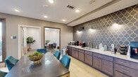 This well-appointed business lounge is thoughtfully designed across a single floor, comprising two distinct functional areas that together create a professional and inviting environment. The main lounge area features large windows dressed with sheer white curtains, allowing abundant natural light to fill the space and offering views of the surrounding greenery. Large-format porcelain tile flooring, warm taupe walls, and a sliding barn door add a modern yet refined aesthetic. Vibrant teal velvet seating is paired with a sleek dark-topped table, bringing a bold pop of color to the space. A standout kitchenette runs along one wall, finished with dark wood cabinetry, light granite countertops, and a striking geometric patterned backsplash in charcoal and gold, accented by wall-mounted sconce lighting. The kitchenette is equipped with a sink and coffee station, making it ideal for hosting or daily use. The adjacent private conference room features neutral walls, carpeted flooring, and natural light through a window with layered curtains. A large rectangular conference table surrounded by quilted leather chairs provides a focused setting for meetings. Potted plants throughout add a welcoming, organic touch to the overall professional ambiance.