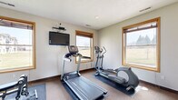 Located in Champlin, MN, this dedicated exercise room offers a compact yet functional fitness space designed to support a well-rounded workout routine. The room is finished with neutral-toned walls, carpeted flooring with protective rubber mats beneath the equipment, and warm wood-trimmed windows that allow ample natural light while offering views of the surrounding green landscape.

The cardio area is equipped with a treadmill and an elliptical trainer, both positioned near the windows to take advantage of the outdoor views during workouts. A wall-mounted flat-screen television is conveniently positioned for entertainment during cardio sessions. On the strength training side, the room houses a multi-station cable weight machine, suitable for a variety of resistance exercises, as well as a set of free weights and a barbell. A full-length mirror with a wood frame is also present, allowing users to monitor their form during exercise.

The room features two interior doors — one with a frosted glass panel serving as the main entry — providing easy access. A small side table adds a practical touch for storing personal items. Ceiling-mounted recessed lighting and ventilation ensure the space remains comfortable during use. This exercise room provides a convenient, self-contained fitness facility well-suited for everyday use.