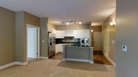 This single-floor residence offers a thoughtfully designed open-concept layout that seamlessly connects the kitchen and living areas. The kitchen is a standout feature, showcasing white cabinetry that extends to the upper walls, complemented by a dark subway tile backsplash and rich dark granite countertops. A central gray island provides additional workspace and serves as a natural focal point for the room. Stainless steel appliances, including a French door refrigerator with an in-door ice and water dispenser and a built-in microwave, add a sleek, modern touch. Track lighting overhead illuminates the kitchen efficiently, while warm-toned walls in a neutral taupe create a cohesive and inviting ambiance throughout. The transition from the kitchen's hardwood-style flooring to plush carpeting in the adjoining living space defines each zone while maintaining an open feel. A hallway visible from the main living area leads to additional rooms, suggesting a well-distributed floor plan. The entryway, visible at the far end of the space, features classic raised-panel door detailing. Overall, the residence balances contemporary finishes with a warm, functional design.