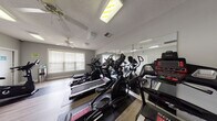 Located at 160 Peaceful Ln in Sanford, NC, this dedicated fitness center is a well-equipped, single-floor space designed to support a comprehensive workout routine. The room features light gray walls, a textured ceiling, and durable hardwood-style flooring with protective equipment mats throughout, creating a clean and functional environment.

The facility is stocked with a variety of cardio equipment, including multiple SOLE-brand treadmills and elliptical machines, as well as upright and cycling stationary bikes. A full-length mirrored wall runs along one side of the room, visually expanding the space and allowing users to monitor their form during workouts. The mirror also reflects the equipment, giving the room an open, spacious feel.

Natural light filters in through multiple windows fitted with horizontal blinds, providing a bright and welcoming atmosphere. Ceiling fans are positioned throughout the space to ensure adequate airflow and comfort during exercise. Overhead fluorescent lighting supplements natural light for consistent illumination at any time of day.

A wall-mounted television is available for entertainment during workouts, and a sanitization station with hand sanitizer is conveniently placed within the room. The space is thoughtfully arranged to allow ease of movement between equipment, making it a practical and accessible fitness facility.