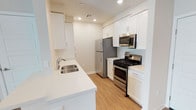 The kitchen is designed with a clean, contemporary aesthetic that maximizes both functionality and visual appeal. White shaker-style cabinetry lines the walls from floor to ceiling, providing generous storage space while contributing to the room's bright, airy atmosphere. Recessed lighting is installed throughout the ceiling, ensuring consistent illumination across the entire workspace.

The countertops are finished in a crisp white surface, offering ample prep space along both sides of the galley-style layout. A stainless steel double-basin sink is positioned along one counter, accompanied by a modern chrome faucet. The appliance suite is entirely stainless steel, including a gas range with oven, an over-the-range microwave, and a full-sized refrigerator — all of which complement the kitchen's polished look.

Light wood-look flooring runs the length of the space, adding warmth and contrast to the predominantly white palette. The galley configuration creates an efficient workflow, with appliances and prep areas thoughtfully arranged on opposing sides. Visible HVAC venting integrated into the ceiling reflects practical attention to comfort and climate control throughout the space.
