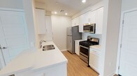 The kitchen is designed with a clean, contemporary aesthetic that maximizes both functionality and visual appeal. White shaker-style cabinetry lines the walls from floor to ceiling, providing generous storage space while contributing to the room's bright, airy atmosphere. Recessed lighting is installed throughout the ceiling, ensuring consistent illumination across the entire workspace.

The countertops are finished in a crisp white surface, offering ample prep space along both sides of the galley-style layout. A stainless steel double-basin sink is positioned along one counter, accompanied by a modern chrome faucet. The appliance suite is entirely stainless steel, including a gas range with oven, an over-the-range microwave, and a full-sized refrigerator — all of which complement the kitchen's polished look.

Light wood-look flooring runs the length of the space, adding warmth and contrast to the predominantly white palette. The galley configuration creates an efficient workflow, with appliances and prep areas thoughtfully arranged on opposing sides. Visible HVAC venting integrated into the ceiling reflects practical attention to comfort and climate control throughout the space.