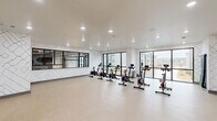 Located at 11420 Bass Pro Pkwy in Little Rock, Arkansas, this exercise room is a purpose-built fitness studio designed for cycling and group workouts. The space is thoughtfully designed with a modern aesthetic, featuring walls adorned with a striking geometric patterned wallcovering in white and black that gives the room a clean, contemporary feel.

A row of professional-grade spin bikes with red and black detailing is arranged across the light wood-style flooring, providing ample space for individual workouts. Large floor-to-ceiling windows line one side of the room, flooding the interior with natural light and offering views of the surrounding community, creating an energizing atmosphere for exercise.

One wall is decorated with an eye-catching arrangement of organically shaped mirrors of varying sizes, which not only serve a functional purpose for form-checking during workouts but also add a distinctive artistic element to the space. A wall-mounted flat-screen television is positioned centrally for entertainment or guided workout viewing.

Recessed ceiling lighting ensures consistent, bright illumination throughout. The open floor plan provides flexibility beyond cycling, with open floor space suitable for stretching or floor exercises. The studio is accessible via multiple entry points, enhancing convenience and flow for users.