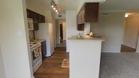 This single-floor residence showcases a well-organized kitchen as its central feature. The kitchen is equipped with dark espresso-toned cabinetry that provides ample storage space, complemented by light-colored countertops, including a peninsula-style breakfast bar that separates the kitchen from the adjoining living area. The flooring throughout the kitchen is a rich, dark hardwood-style material that adds warmth and contrast to the neutral gray walls. Appliances include a white refrigerator, an electric range, and a built-in microwave, all positioned along a single functional wall. Track lighting runs along the ceiling, providing direct illumination over the cooking and prep areas. A ceiling vent ensures proper climate control. The kitchen opens through a corridor-style layout toward additional rooms, with a glimpse of a bathroom visible in the background. As the space transitions away from the kitchen, the flooring shifts to carpet, suggesting a comfortable living or bedroom area beyond. The overall design is practical and cohesive, with consistent cabinetry tones and neutral wall colors throughout.