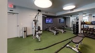 Located at 30 W Howell St in Boston, MA, this well-appointed fitness center offers a comprehensive and thoughtfully designed workout environment across a single level. The space is characterized by a contemporary aesthetic, featuring dark accent walls, a blue-grey ceiling, and large circular overhead lighting fixtures that provide bright, even illumination throughout.

The cardio zone benefits from generous natural light streaming in through floor-to-ceiling windows that overlook the surrounding outdoor area, creating an open and energizing atmosphere. This section houses multiple treadmills, elliptical trainers, and a stationary bike, all equipped with individual display screens.

A dedicated functional training area features artificial turf flooring and includes rowing machines, battle ropes, a cable machine, TRX suspension straps, and a multi-functional trainer. Wall-mounted flat-screen TVs enhance the workout experience in this zone, which also features full-length mirrors to maximize the sense of space.

The strength training section is equally well-equipped, with a power rack, adjustable benches, a comprehensive dumbbell rack, barbell sets with weight plates, and medicine balls. The combination of dark rubberized flooring and green turf throughout the space reinforces its professional, gym-quality environment. The fitness center provides residents with a versatile, full-body training facility designed to meet a wide range of fitness needs.