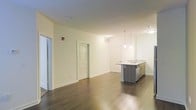 This single-floor space features a clean, contemporary design with an open-concept layout that seamlessly connects the living area and kitchen. Rich, dark hardwood flooring extends throughout, providing a warm contrast to the bright white walls and trim. The kitchen is anchored by a sleek island with a stone countertop and integrated sink, complemented by modern gray cabinetry and stainless steel appliances. Elegant pendant lights hang above the island, adding a refined touch to the space. Recessed and flush-mount ceiling lighting ensures the entire area is well-illuminated, enhancing the sense of openness. The generous living area offers ample room for flexible furniture arrangements, while multiple interior doors suggest well-defined private rooms beyond the main living space. Crisp white panel doors and wide baseboards throughout contribute to the polished, modern aesthetic. The overall ambiance is bright, airy, and move-in ready, making it a versatile and functional space suited to a variety of living arrangements.