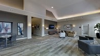 This single-floor space presents a well-appointed, open-concept layout characterized by soaring vaulted and tray ceilings that amplify the sense of volume throughout the interior. The flooring features a patterned, multi-toned carpet in earthy gray and beige tones that anchors the space and adds textural interest underfoot. Structural columns define distinct zones without fully enclosing them, maintaining an open and airy flow between areas.

The main seating area is furnished with a neutral-toned sofa, an upholstered armchair, and a side table, arranged around a wall-mounted flat-screen television positioned above a decorative geometric credenza. Wall-mounted sconces and floor lamps provide warm, layered lighting that complements the recessed ceiling fixtures. Curated artwork adorns the walls, contributing to a refined, contemporary aesthetic.

A connected corridor visible through an open column framing leads further into the space, lined with additional framed artwork and wall sconces. The overall ambiance is polished and welcoming, blending modern design elements with comfortable furnishings to create a functional yet sophisticated communal environment.