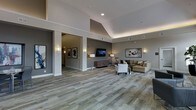 This single-floor space presents a well-appointed, open-concept layout characterized by soaring vaulted and tray ceilings that amplify the sense of volume throughout the interior. The flooring features a patterned, multi-toned carpet in earthy gray and beige tones that anchors the space and adds textural interest underfoot. Structural columns define distinct zones without fully enclosing them, maintaining an open and airy flow between areas.

The main seating area is furnished with a neutral-toned sofa, an upholstered armchair, and a side table, arranged around a wall-mounted flat-screen television positioned above a decorative geometric credenza. Wall-mounted sconces and floor lamps provide warm, layered lighting that complements the recessed ceiling fixtures. Curated artwork adorns the walls, contributing to a refined, contemporary aesthetic.

A connected corridor visible through an open column framing leads further into the space, lined with additional framed artwork and wall sconces. The overall ambiance is polished and welcoming, blending modern design elements with comfortable furnishings to create a functional yet sophisticated communal environment.