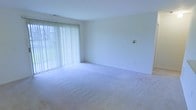 This single-floor space presents a clean, open layout finished with wall-to-wall light-toned carpeting that extends throughout the room, lending a sense of warmth and continuity underfoot. The walls are painted in a crisp, neutral white, contributing to the overall brightness and airy feel of the interior. A defining feature of the space is the large sliding glass door fitted with full-length vertical blinds, which spans a generous portion of one wall. This architectural element allows an abundance of natural light to filter into the room, illuminating the space evenly throughout the day while also providing direct access to an outdoor area with visible greenery beyond. The ceiling is standard height and smooth, maintaining a simple, unadorned aesthetic that keeps the focus on the open floor plan. A light switch plate is visible on the adjacent wall, and a baseboard electrical outlet provides practical utility. The overall ambiance is understated and versatile, offering a neutral canvas well-suited to a variety of interior arrangements and décor styles. The combination of natural light, soft carpeting, and clean white walls creates a comfortable, move-in-ready atmosphere.