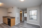 This single-floor unit offers a practical and cohesive layout designed for everyday comfort. The kitchen serves as a central feature, equipped with black appliances including an electric range, refrigerator, and dishwasher. Light maple cabinetry pairs with dark granite-style countertops, providing ample storage and workspace. A stainless steel double sink sits along one wall, and light wood-look flooring runs throughout the kitchen and adjoining areas, contributing to a clean, consistent aesthetic.

The open layout connects the kitchen to the living area, which transitions to soft carpeting underfoot. A ceiling fan with an integrated light fixture provides both air circulation and ambient lighting in this space. Natural light enters through windows fitted with blinds, adding brightness to the interior.

The bathroom features the same warm cabinetry and dark countertop found in the kitchen, topped with a white basin sink. A large mirror, towel bar, and wood-look flooring complete the well-appointed space.

The bedroom is a generously sized, carpeted room with neutral walls and a ceiling fan, offering a quiet and comfortable retreat. Overall, the unit maintains a consistent, neutral palette throughout, creating a cohesive and functional living environment.