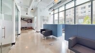 Located at 125 NE 32nd St in Miami, FL, this single-floor commercial space offers a thoughtfully designed open-plan environment that balances collaborative and private work settings. The layout features polished concrete terrazzo flooring throughout, lending a sleek, professional aesthetic that complements the industrial-chic exposed ceiling with visible ductwork, conduit, and pendant lighting fixtures.

The main open area is anchored by modular lounge seating in deep charcoal and muted blue tones, arranged around freestanding blue booth partitions that create semi-private nooks ideal for focused work or informal meetings. Floor-to-ceiling glass walls enclose a dedicated conference or private office area, maintaining visual openness while providing acoustic separation.

Abundant natural light streams in through expansive windows along the perimeter, offering views of the surrounding Miami streetscape and lush greenery. The high ceilings amplify the sense of spaciousness throughout.

A standout feature is the kitchenette area, which boasts a geometric slatted wood accent wall adorned with a glowing neon "Today's Vibe" sign, paired with clean white cabinetry and a beverage refrigerator — adding a vibrant, contemporary personality to the space. The overall design strikes a balance between modern functionality and creative energy, making it well-suited for a dynamic professional environment.