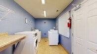 This shared laundry facility is a clean, efficiently organized space designed to meet the practical needs of its users. The room is finished with distinctive periwinkle-blue walls and light tile flooring, giving it a bright and tidy appearance. Overhead fluorescent lighting ensures the space is well-lit throughout.

The facility is outfitted with multiple front-load and top-load washing machines, as well as a row of dryers, providing ample capacity for simultaneous use. A built-in granite-style folding counter runs along one wall, offering a convenient surface for sorting and folding laundry. An open wooden shelving unit beneath the counter provides additional storage space for laundry supplies.

A built-in L-shaped bench seat is positioned near a window dressed with vertical blinds, which allows natural light to filter in and provides a pleasant view of the surrounding greenery — a comfortable spot to wait while laundry cycles complete. Wire shelving mounted on the wall above offers further organizational utility.

Safety is accounted for with a prominently mounted fire extinguisher near the entry door. Informational signage is posted on the walls, and the space is equipped with ventilation registers. The single-floor layout is straightforward and accessible, making this laundry room a practical and functional shared amenity.