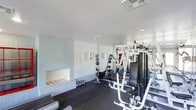 This single-floor fitness space is thoughtfully designed to support a comprehensive workout experience. The room features painted white brick walls that lend an industrial yet clean aesthetic, complemented by a smooth white ceiling with recessed and flush-mounted lighting that keeps the space bright and functional. A large mirrored wall along one side of the room visually expands the space while allowing users to monitor their form during exercise. The flooring consists of durable rubber matting, ideal for heavy equipment and high-impact training. On one side of the room, a built-in fireplace-style niche is set into the brick wall, adding a distinctive architectural detail. The space is outfitted with a multi-station cable and weight machine, an adjustable bench, a rack of kettlebells, and a medicine ball storage rack offering a variety of weighted balls. Wall-mounted accessories, such as agility ladders, are also present. The overall ambiance is clean, practical, and purpose-driven, making this a well-rounded fitness facility suited for both strength training and functional fitness routines.