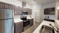 This single-floor residence offers a thoughtfully designed open-concept layout that seamlessly connects the kitchen and living areas. The kitchen is a standout feature, showcasing dark espresso-toned cabinetry complemented by lighter upper cabinets, bright white countertops, and a full suite of stainless steel appliances including a refrigerator, electric range, and built-in microwave. A spacious kitchen island with an integrated sink provides both additional prep space and a natural gathering point. Pendant lighting and a track lighting system overhead add both function and style to the cooking space. A dedicated walk-in pantry offers generous storage directly off the kitchen. Wood-style plank flooring runs throughout the main living areas, lending a warm, cohesive feel, while soft carpet adds comfort to the bedroom, which includes a closet with built-in shelving. The bathroom features a combination tub and shower, a single-sink vanity with dark cabinetry, and wood-style flooring that ties the space together. A dedicated laundry room houses a full-size washer and dryer with wire shelving above for added storage. The open living area is generously sized, filled with natural light, and provides a versatile, move-in-ready space throughout.
