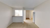 This single-floor residential space offers a straightforward, functional layout designed for comfortable everyday living. The living area is finished with wall-to-wall carpet in a neutral tone, creating a warm and understated atmosphere throughout. A defining architectural feature is the pass-through kitchen window, which opens the kitchen to the main living space, allowing for easy interaction between the two areas while maintaining a sense of separation. The kitchen is outfitted with white cabinetry and appliances, a countertop with a built-in sink, and a serving ledge that doubles as a casual dining surface. Adjacent to the kitchen, a structural column adds a subtle visual break without compromising the open feel of the space. Looking toward the entryway, a built-in shelving unit provides convenient storage near the front door, while a tall utility panel is integrated neatly into the wall. The consistent use of light, neutral tones on the walls and ceilings throughout contributes to a clean, cohesive aesthetic. The layout flows efficiently from the living area through to the entry corridor, making the space feel connected and easy to navigate.
