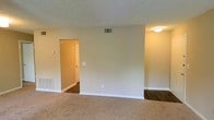 This single-story space presents a clean, functional layout visible through its open living area. The main room features wall-to-wall carpet in a warm neutral tone, complemented by crisp white baseboards and beige walls that create a bright, understated atmosphere. A textured popcorn ceiling adds a classic finish overhead, while a flush-mount ceiling light provides consistent ambient illumination. Ventilation is well-considered, with visible HVAC registers on both the wall and floor, ensuring effective climate control throughout. A doorway leads to an adjoining area finished with dark hardwood-style flooring, offering a distinct contrast in materials. This secondary space features a louvered closet door, suggesting a utility or storage area. A wall-mounted thermostat is conveniently positioned near the transition between rooms, allowing easy access to climate settings. The overall design is straightforward and practical, with a neutral color palette throughout that offers a versatile backdrop suitable for a variety of furnishing styles. The single-floor layout promotes ease of navigation and accessibility throughout the space.