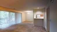 This single-floor residence offers a thoughtfully designed open-concept layout that seamlessly connects the main living and dining areas. The living room is generously proportioned, featuring soft wall-to-wall carpeting underfoot and a large sliding glass door fitted with vertical blinds, which allows an abundance of natural light to filter into the space while providing access to the outdoors. The dining area is distinguished by an elegant chandelier that adds a refined touch to the otherwise understated aesthetic. A partial wall divides the living and dining spaces from the kitchen, which features a pass-through counter that maintains visual connectivity throughout the open floor plan. The kitchen is equipped with wood-tone cabinetry and a white refrigerator, providing functional storage and workspace. Recessed lighting in the kitchen area complements the chandelier in the dining space, ensuring consistent illumination throughout. The neutral, muted color palette of warm beige tones on the walls and carpeting creates a calm and cohesive ambiance. The straightforward single-floor layout offers ease of navigation and a comfortable, livable feel throughout the residence.