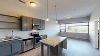 Located at 161 Concord Exchange N in South Saint Paul, MN, this single-floor unit offers a well-designed, contemporary living space with thoughtful finishes throughout. The kitchen is a standout feature, showcasing gray shaker-style cabinetry, a classic herringbone tile backsplash, and a full suite of stainless steel appliances including a refrigerator, electric range, microwave, and dishwasher. A central island with striking marble-patterned countertops provides additional prep space and seating, while pendant lighting adds a warm, polished ambiance. Light wood-look flooring runs through the main living and kitchen areas, lending a clean, cohesive aesthetic. The open layout connects the kitchen to a generously sized living area, which benefits from large windows fitted with horizontal blinds that invite natural light and outdoor views of the surrounding greenery. A carpeted bedroom features its own window with tree-line views, creating a quiet, comfortable retreat. Built-in shelving in a dedicated storage closet offers practical organization space. The bathroom is cleanly appointed with a soaking tub and shower combo, gray vanity cabinetry, white countertops, and wood-look tile flooring, complemented by grab bars for added accessibility. The overall design balances modern style with functional living.