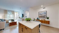 This single-floor residence showcases a thoughtfully designed open-concept layout with modern finishes and a cohesive coastal-inspired aesthetic. The kitchen is a standout feature, equipped with warm wood cabinetry, stainless steel appliances, a striking marble-veined quartz backsplash, and a central island topped with matching quartz that doubles as a casual dining space with seating for two. Light wood-tone flooring flows throughout, enhancing the bright, airy atmosphere created by recessed lighting and white walls.

The living room makes a bold statement with a deep navy accent wall serving as a gallery backdrop, contrasting elegantly with neutral furnishings. Sheer linen curtains frame a sliding glass door that opens to a private balcony, inviting natural light and outdoor views into the space.

The bedrooms are well-appointed and serene. The primary bedroom features a textured geometric accent wall, light wood flooring, and large windows with layered sheer curtains that allow abundant natural light. A secondary bedroom offers clean lines and outdoor views. The bathroom is finished with large-format neutral floor tiles, white subway-tiled walls, matte black fixtures, and a full soaking tub with shower.